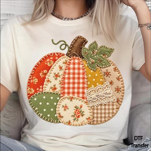 May include: A cream-colored t-shirt featuring a large, central graphic of a patchwork pumpkin. The pumpkin is composed of various fabric patterns including orange floral, red and white gingham, green polka dots, and lace, all outlined with a faux blanket stitch.