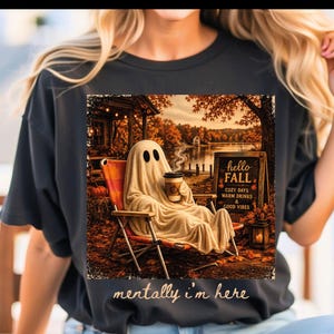 May include: A black t-shirt featuring a graphic of a ghost sitting in a lawn chair holding a coffee cup near a lake. The graphic includes a sign that reads hello FALL COZY DAYS WARM DRINKS & GOOD VIBES and the text mentally i'm here.