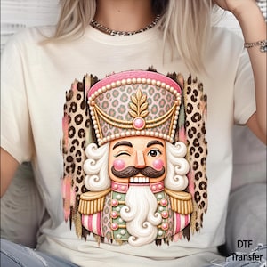 May include: A cream-colored t-shirt featuring a graphic of a winking nutcracker with a pink and green leopard print hat and uniform. The nutcracker has a white beard, mustache, and gold epaulets, set against a brown leopard print brushstroke background.