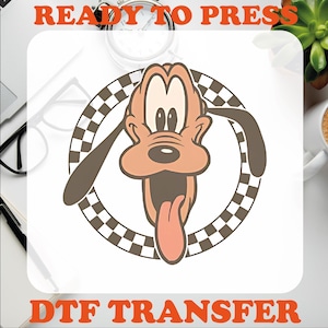 May include: A cartoon image of a brown and orange dog with a checkered background. The dog has a large tongue sticking out and is smiling. The text "READY TO PRESS" is at the top of the image and "DTF TRANSFER" is at the bottom.