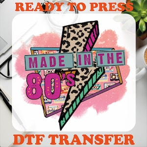 May include: A DTF transfer design featuring a lightning bolt with a leopard print pattern. The design includes the text "MADE IN THE 80'S" in a retro font, set against a pink and cassette tape background. The words "READY TO PRESS" and "DTF TRANSFER" are also included.