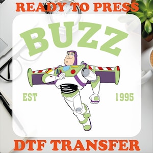 May include: A green, white, and purple cartoon image of Buzz Lightyear from the Toy Story franchise. The image includes the text "Buzz" in a green font, "EST 1995" in a black font, and "READY TO PRESS DTF TRANSFER" in an orange font.