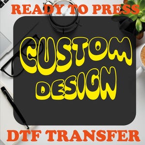 May include: A black background with the text "READY TO PRESS" in orange at the top. The text "CUSTOM DESIGN" is in yellow bubble letters. The text "DTF TRANSFER" is in orange at the bottom.
