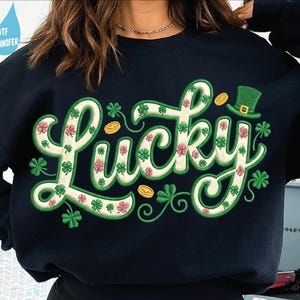 May include: Navy blue sweatshirt with the word "Lucky" in a whimsical, embroidered design. The word is filled with shamrocks and pink flowers, with a green outline. Gold coins and a green leprechaun hat are also featured.