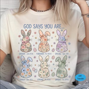 May include: Cream-colored t-shirt featuring a colorful graphic of six Easter bunny illustrations with floral patterns and bows. The text "GOD SAYS YOU ARE..." is above the bunnies, with inspirational words and Bible verses below each bunny.