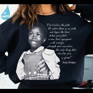 May include: Black sweatshirt featuring a black and white image of Ruby Bridges and a quote. The quote reads: "Don't follow the path. Go where there is no path and begin the trail..." -Ruby Bridges. A DTF transfer is visible in the upper left corner.