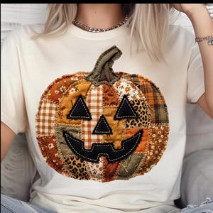 May include: A cream-colored t-shirt featuring a large, central graphic of a pumpkin made from a patchwork of various fabrics. The pumpkin design includes brown, orange, and green patterns, including plaid, floral, and leopard print, with black fabric eyes, nose, and a jagged mouth.