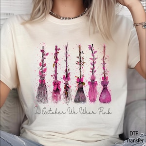 May include: A cream-colored t-shirt featuring a graphic of six stylized broomsticks decorated with pink leaves and bows. Below the broomsticks, the text reads In October We Wear Pink. The design includes pink paint splatter accents.