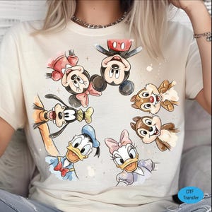 May include: Cream-colored t-shirt featuring a watercolor design of Mickey Mouse, Minnie Mouse, Donald Duck, Daisy Duck, Goofy, Chip, and Dale. The characters are arranged in a circle. The shirt has a DTF Transfer label.