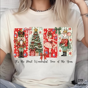 May include: A cream-colored t-shirt featuring a graphic print of a nutcracker, a Christmas tree, a ballerina, and a mouse in a crown. The text "It's the Most Wonderful Time of the Year" is printed below the illustrations in a black script font.