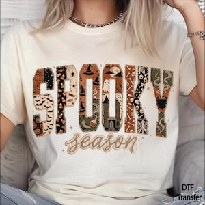 May include: A cream-colored t-shirt featuring the word SPOOKY in large, blocky letters filled with various Halloween patterns including pumpkins, bats, ghosts, and a haunted house. Below, the word season is written in a cursive script font.