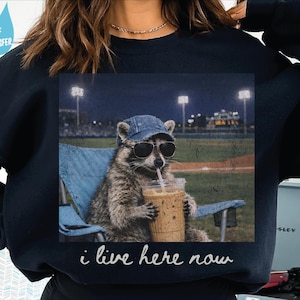 May include: Navy blue sweatshirt featuring a raccoon wearing sunglasses and a denim cap, drinking from a clear cup with a straw. The raccoon is sitting in a blue chair with a baseball field background. The text "i live here now" is below the image.