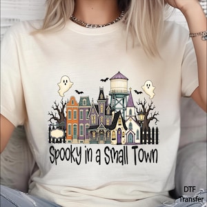 May include: A cream-colored short-sleeve t-shirt featuring a graphic of a small town with colorful buildings, a water tower, bare trees, and two ghosts. The text "Spooky in a Small Town" is printed in black below the illustration of the town.