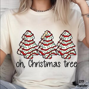 May include: A cream-colored t-shirt featuring a graphic of three Christmas tree-shaped cookies with white icing, red drizzle, and green sprinkles. The middle cookie has a bite taken out of the top. The text "oh, Christmas tree" is printed below.