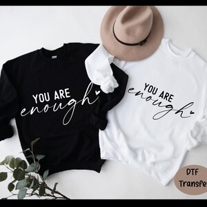 May include: Two sweatshirts, one black and one white, each with the phrase "YOU ARE enough" in a cursive font. A tan hat rests on top of the white sweatshirt. The black sweatshirt has a small heart next to the word "enough".