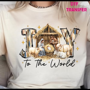 May include: Cream-colored t-shirt featuring a nativity scene with the words "JOY To The World." The word "JOY" is in a patterned font, and the scene includes Mary, Joseph, baby Jesus, sheep, and a stable.