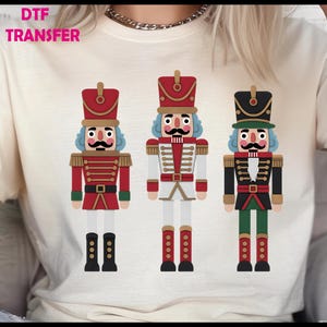 May include: A cream-colored t-shirt featuring three colorful nutcracker soldier designs. The nutcrackers have red, white, and black uniforms with gold accents, and each has a unique hat and facial expression. The text "DTF TRANSFER" is at the top left.