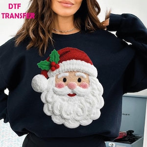 May include: Navy blue sweatshirt featuring a detailed Santa Claus design. The Santa Claus graphic includes a red hat, white beard, and rosy cheeks. The text "DTF TRANSFER" is visible in the upper left corner.