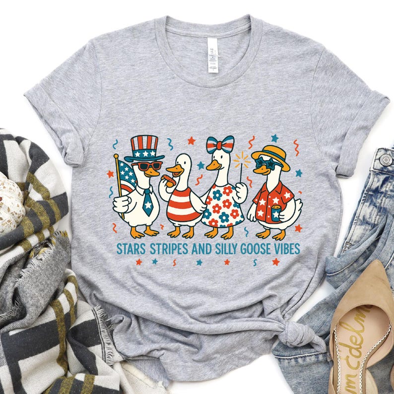May include: A light gray t-shirt featuring a whimsical illustration of four cartoon geese dressed in patriotic attire. The geese are wearing sunglasses, hats, and holding a flag, sparkler, and drink. The text "STARS STRIPES AND SILLY GOOSE VIBES" is below.