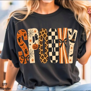 May include: A black t-shirt featuring the word SPOOKY in large, stylized letters. Each letter has a unique pattern: orange with ghosts and bats, cream with spiderwebs and a pumpkin, leopard print, black and white checkers, orange stripes with a bow, and cream with stars and a witch hat.