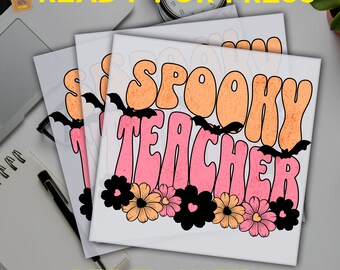 May include: Stacked white DTF transfer sheets with the words "SPOOKY TEACHER" in orange and pink, decorated with black bats and flowers. The words "READY FOR PRESS" and "DTF TRANSFERS" are also visible.