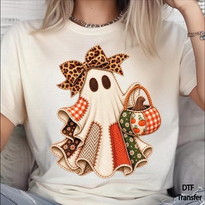 May include: A cream-colored t-shirt featuring a graphic of a ghost wearing a leopard print bow. The ghost is decorated with various fabric-like patches in orange, brown, and green patterns, and it is holding a pumpkin-filled basket.