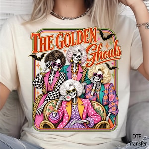May include: A cream-colored t-shirt featuring a graphic print of four skeletons with blonde hair, wearing colorful patterned blazers and sunglasses, sitting on a sofa. The text "THE GOLDEN Ghouls" is printed in orange above the skeletons, with small black bats and star accents.