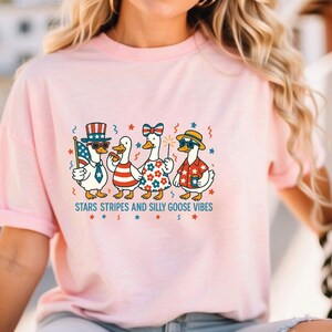 May include: Pink short-sleeved t-shirt featuring a graphic of four cartoon geese dressed in patriotic attire. The geese are wearing hats, sunglasses, and holding a flag and sparkler. The text "STARS STRIPES AND SILLY GOOSE VIBES" is printed below the geese.