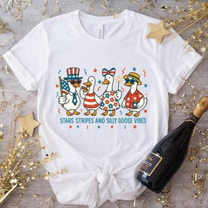 May include: White t-shirt featuring a cartoon illustration of four geese dressed in patriotic attire, including sunglasses, hats, and a bow. The text "STARS STRIPES AND SILLY GOOSE VIBES" is printed below the geese. A bottle of Prosecco is in the background.