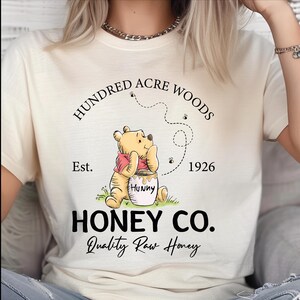 May include: A cream-colored t-shirt featuring a graphic of Winnie the Pooh with a honey pot. The text reads HUNDRED ACRE WOODS, Est. 1926, HONEY CO., and Quality Raw Honey. Small bees and a dotted flight path are printed above the character.