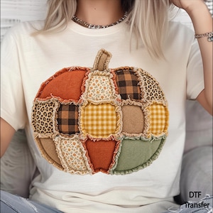 May include: A cream-colored short-sleeve t-shirt featuring a large, central graphic of a patchwork pumpkin. The pumpkin design is composed of various fabric textures including orange, olive green, brown plaid, yellow gingham, leopard print, and floral patterns.