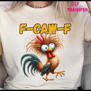 May include: A cream-colored t-shirt featuring a cartoon rooster with wide eyes and a red comb. The rooster has brown and orange feathers and a blue and green tail. The text "F-CAW-F" is printed above the rooster in yellow letters with a black outline.