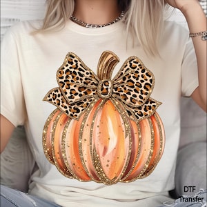 May include: A cream-colored t-shirt featuring a large graphic of an orange pumpkin with gold glitter accents and a leopard print bow on top. The text DTF Transfer is printed in the bottom right corner of the shirt design.