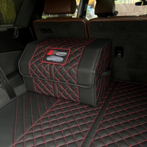 May include: Black car trunk organizer with red diamond stitching. The organizer is rectangular with a red and black logo. The trunk mat also has red diamond stitching. The car interior has brown leather seats.