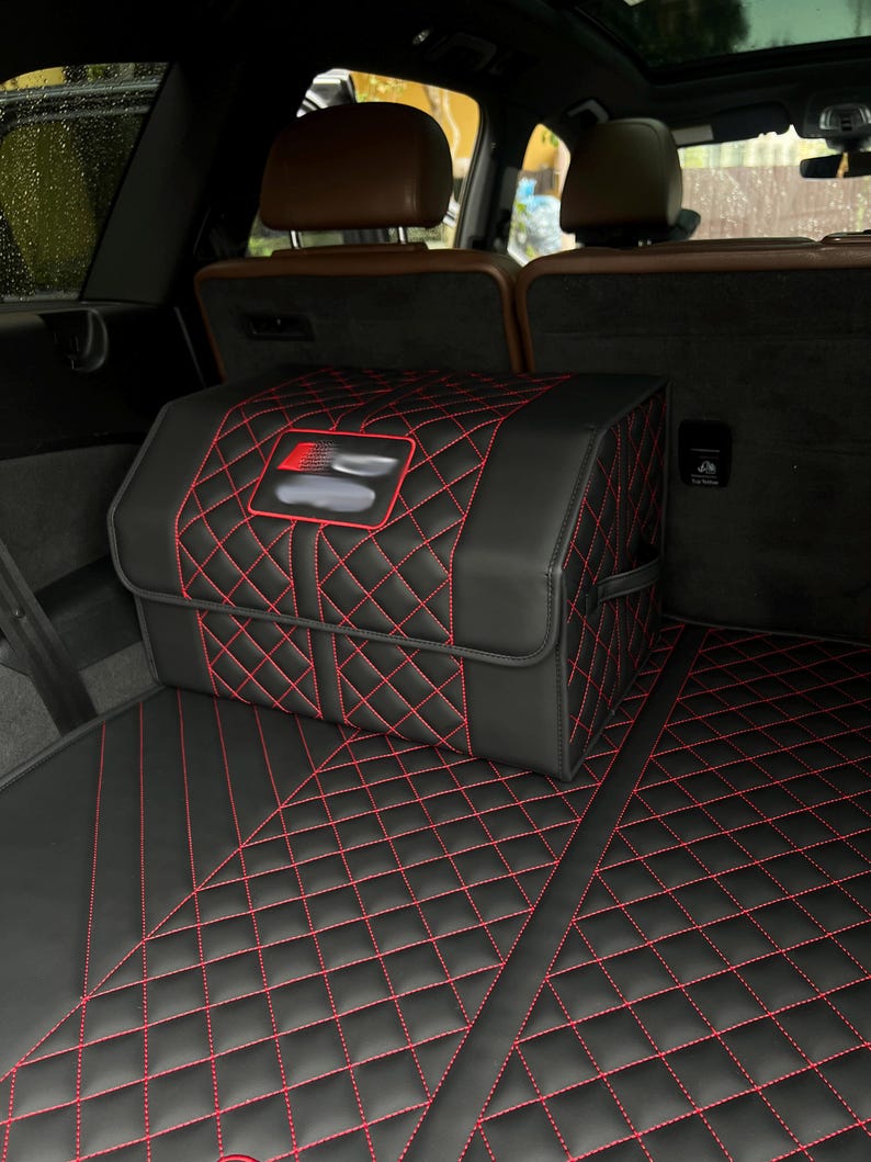 May include: Black car trunk organizer with red diamond stitching. The organizer is rectangular with a red and black logo. The trunk mat also has red diamond stitching. The car interior has brown leather seats.