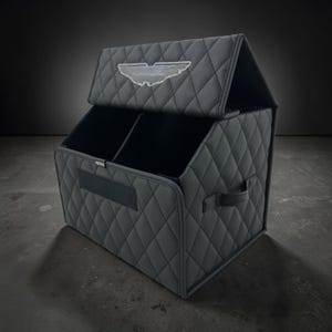 May include: A black, quilted storage organizer with a divided interior and a hinged lid. The organizer features a diamond pattern, a handle, and a logo on the lid. The organizer is designed for car storage.