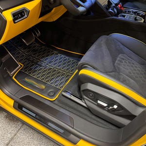 May include: Interior view of a luxury car with a yellow and black color scheme. The seats are black with yellow accents, and the floor mats have a geometric pattern. The dashboard and door panels are yellow, and the steering wheel is black.