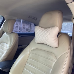 May include: Beige leather car seat with a white quilted headrest pillow. The seat has a horizontal stitched pattern.