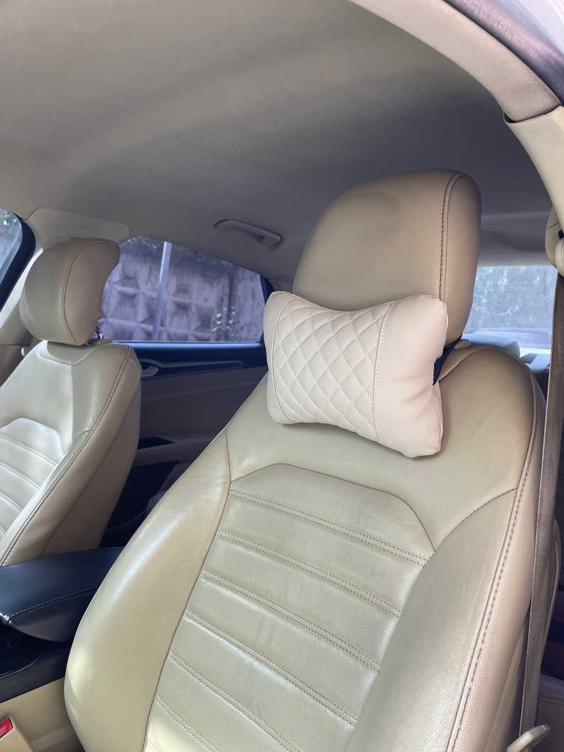 May include: Beige leather car seat with a white quilted headrest pillow. The seat has a horizontal stitched pattern.