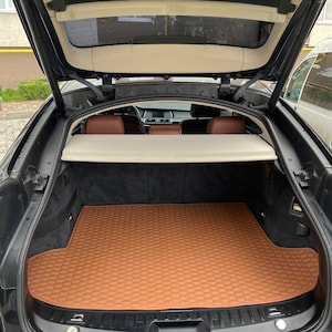 Puede incluir: Maletero abierto de un coche negro, que revela una alfombra marrón texturizada que cubre el suelo. El interior presenta asientos de cuero marrón y un revestimiento de techo beige. El maletero está abierto, mostrando el espacio de almacenamiento del coche.