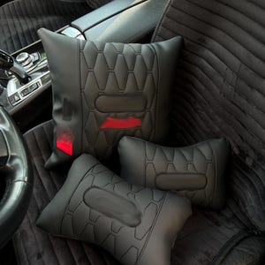 May include: Three black car seat pillows with a quilted geometric pattern. The pillows are arranged on a black car seat. One pillow has a red accent. The pillows appear to be made of leather or a similar material.