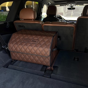 Puede incluir: Una caja de almacenamiento acolchada marrón con un asa negra en la parte trasera de un coche. La caja está situada en el suelo del coche, detrás de los asientos traseros.