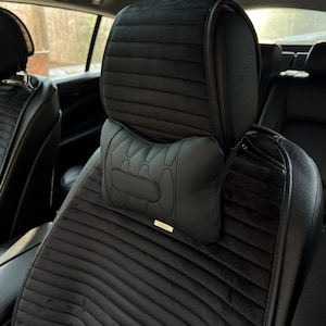 May include: Black car seat with a headrest and a neck pillow. The seat has a ribbed, velvety texture. The headrest and neck pillow are black leather. The neck pillow has a stitched design and a small gold-colored accent.