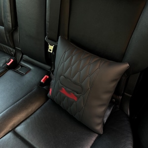 May include: Black car seat headrest pillow with a geometric pattern and red accents. The pillow is secured by a seat belt and is designed to provide comfort and support while driving.