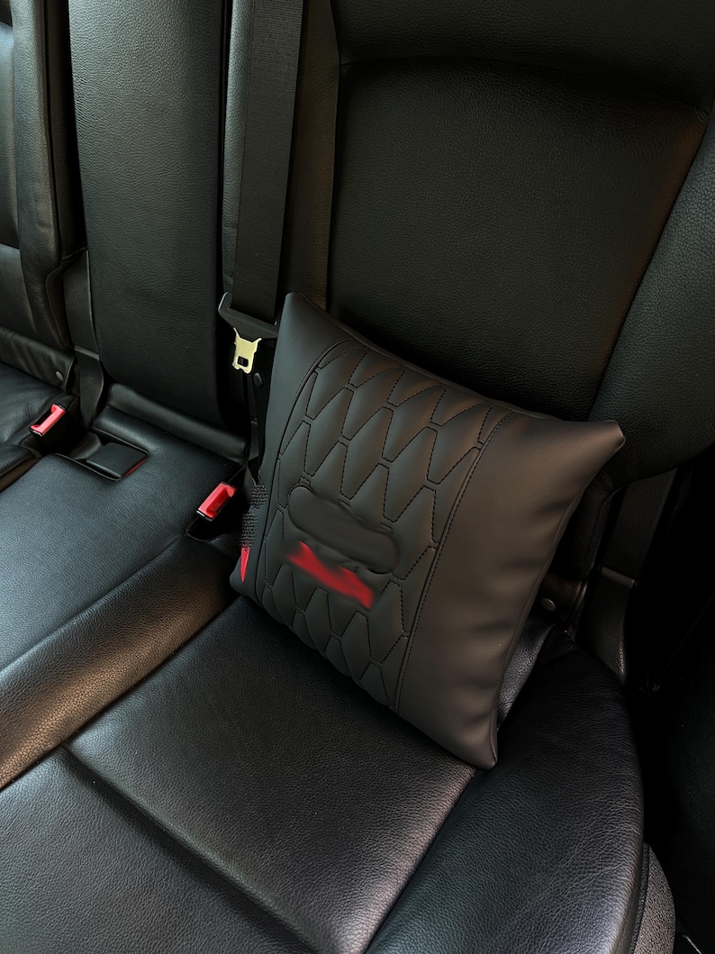 May include: Black car seat headrest pillow with a geometric pattern and red accents. The pillow is secured by a seat belt and is designed to provide comfort and support while driving.
