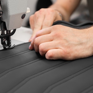 May include: A close-up of a person's hands sewing black leather with a white industrial sewing machine. The leather has a quilted pattern.
