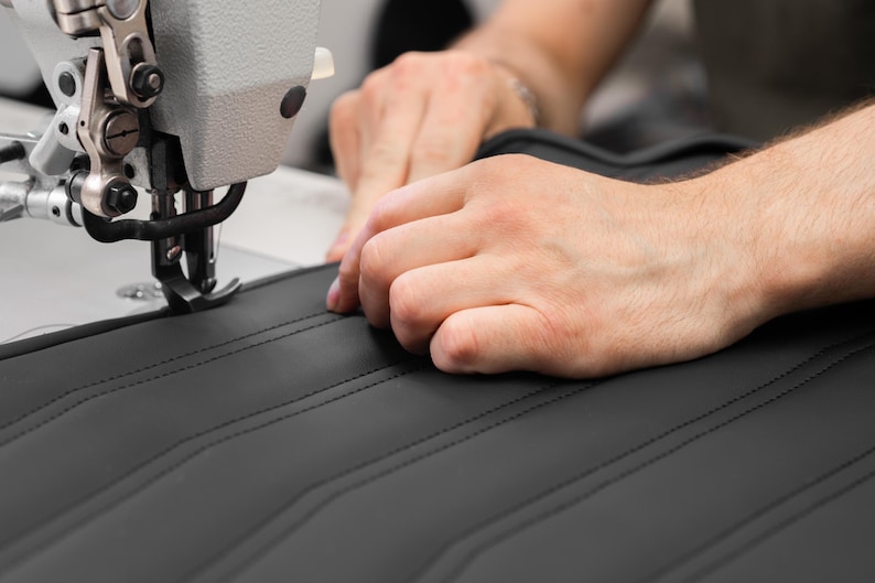 May include: A close-up of a person's hands sewing black leather with a white industrial sewing machine. The leather has a quilted pattern.
