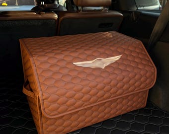 May include: A brown, quilted car organizer with a white emblem on the front. The organizer is rectangular with a folding lid and handles. The interior of the car is visible in the background.