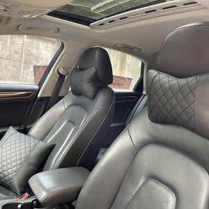 May include: Two black quilted headrest pillows for car seats. The pillows are in the front seats of a black leather interior car.