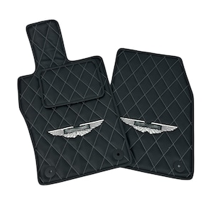 May include: Black car floor mats with a diamond-stitched pattern and white stitching. Each mat features a silver emblem with a wing design. The set includes a driver-side mat, a passenger-side mat, and a smaller mat.